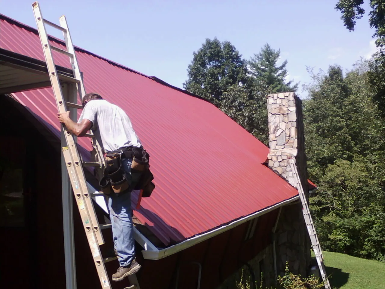 Roofer on a ladder beside a red metal roof and stone chimney — Soffit & Fascia Repair by Roofing Brothers in Oneida